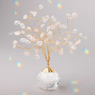 A decorative crystal tree centerpiece with gold-tone twisted branches, clear faceted crystal drops, and pale blush pink stone leaves, mounted on a clear faceted crystal sphere base against a soft gray background with subtle rainbow light leaks.