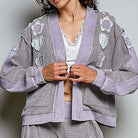 A model wearing a lavender and gray houndstooth cardigan with floral appliques and matching pants, styled over a white lace-trimmed top against a neutral gray background.
