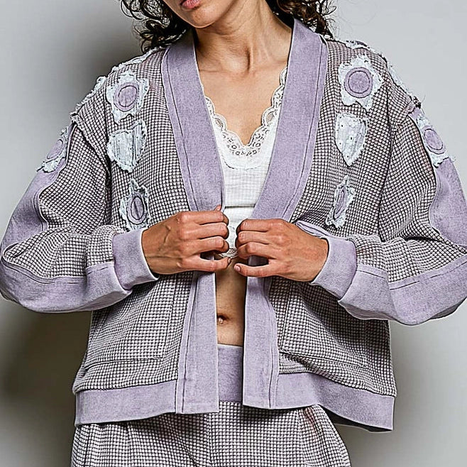 A model wearing a lavender and gray houndstooth cardigan with floral appliques and matching pants, styled over a white lace-trimmed top against a neutral gray background.