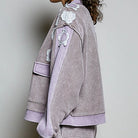A model shown in profile wearing a lavender and gray houndstooth jacket with lavender ribbed trim and white floral appliques, paired with matching houndstooth pants against a light gray studio background.