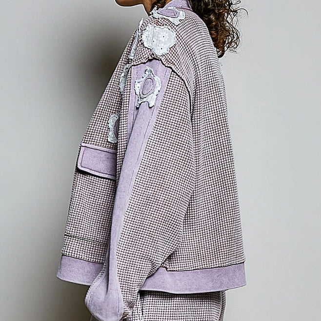 A model shown in profile wearing a lavender and gray houndstooth jacket with lavender ribbed trim and white floral appliques, paired with matching houndstooth pants against a light gray studio background.