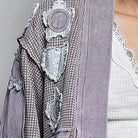 A close-up of a model wearing a textured lavender-gray patchwork cardigan over a white lace-trimmed top, against a neutral gray background.