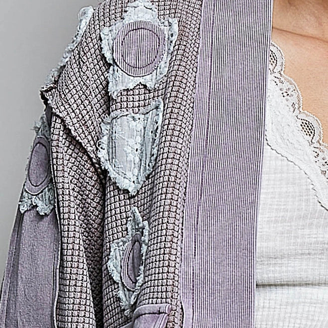 A close-up of a model wearing a textured lavender-gray patchwork cardigan over a white lace-trimmed top, against a neutral gray background.