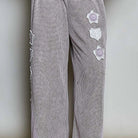 A person wearing loose, wide-leg trousers in a small-scale gray and white checkered pattern with light purple side panels and white embroidered floral details. The person is also wearing black flip-flops and standing on a woven rug against a plain wall.