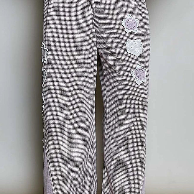A person wearing loose, wide-leg trousers in a small-scale gray and white checkered pattern with light purple side panels and white embroidered floral details. The person is also wearing black flip-flops and standing on a woven rug against a plain wall.