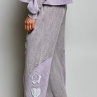 A person wearing wide-leg lounge pants in a light gray and white houndstooth pattern with lavender-colored contrast panels and white embroidered floral details down the leg, paired with black flip-flops.
