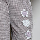 Close-up of a pair of gray and white micro-check patterned trousers featuring three lavender embroidered flower appliqués on the leg, with a matching lavender waistband visible at the top.