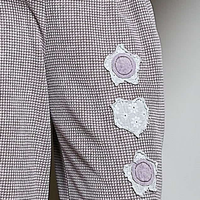 Close-up of a pair of gray and white micro-check patterned trousers featuring three lavender embroidered flower appliqués on the leg, with a matching lavender waistband visible at the top.