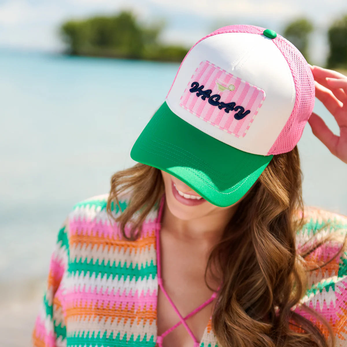 Woman wearing a colorful striped cover-up and a pink and green cap with "Vacay"text, standing by a body of water.